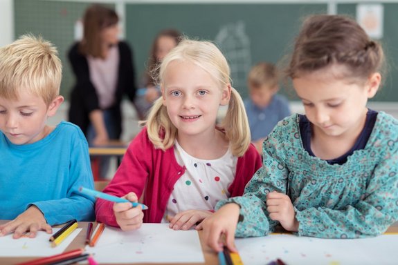 Zwei Mädchen und ein Junge im Grundschulalter vor Heften sitzend mit Stiften in der Hand