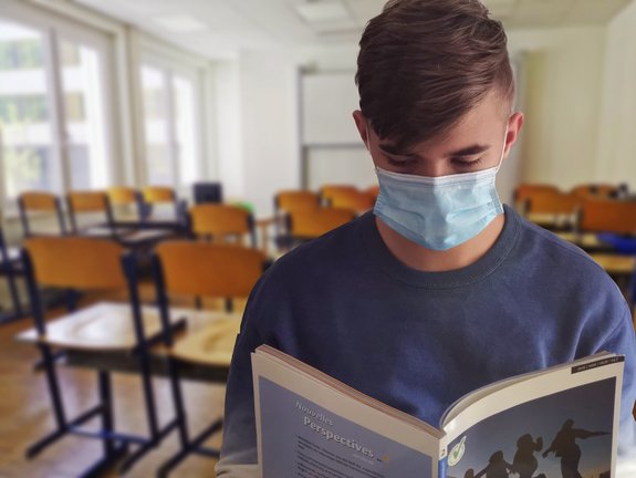 Junge stehend im Klassenzimmer mit Maske ließt ein Buch