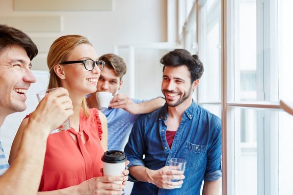 Gruppe junger Männer und Frauen mit Kaffeetassen lachend vor einer Fensterfront