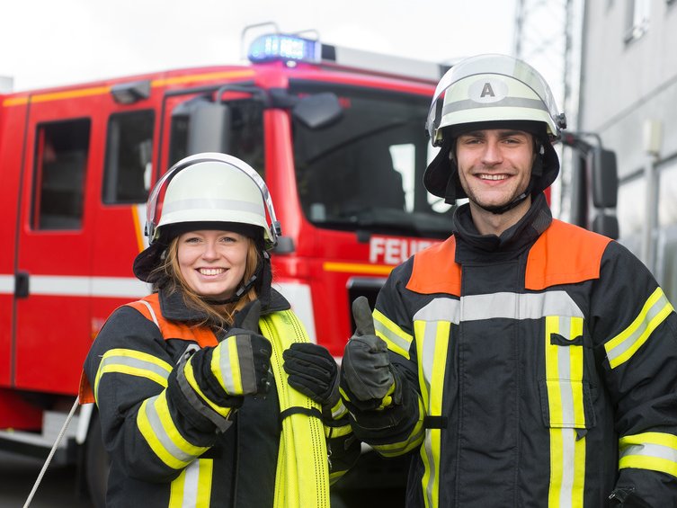 Feuerwehrfrau und Feuerwehrmann vor Einsatzfahrzeug mit nach oben gerichteten Daumen