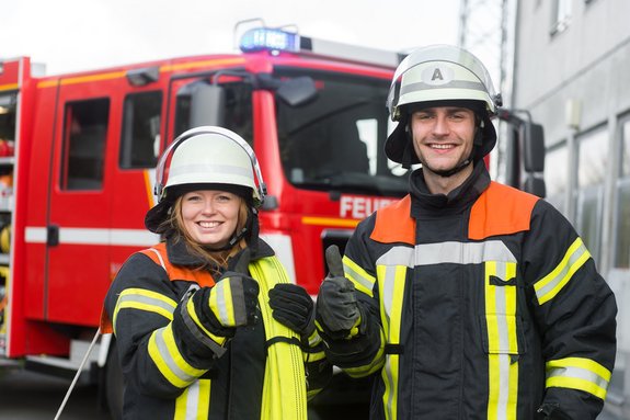 Feuerwehrfrau und Feuerwehrmann vor Einsatzfahrzeug mit nach oben gerichteten Daumen
